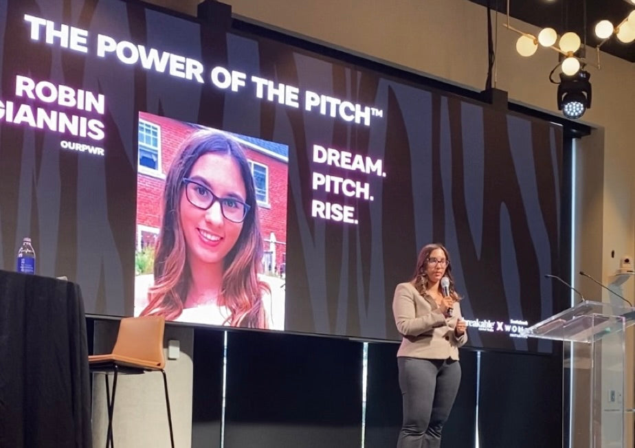 Robin Petrogiannis speaking on stage with a microphone, beside a screen reading The Power of the Pitch — Dream. Pitch. Rise.