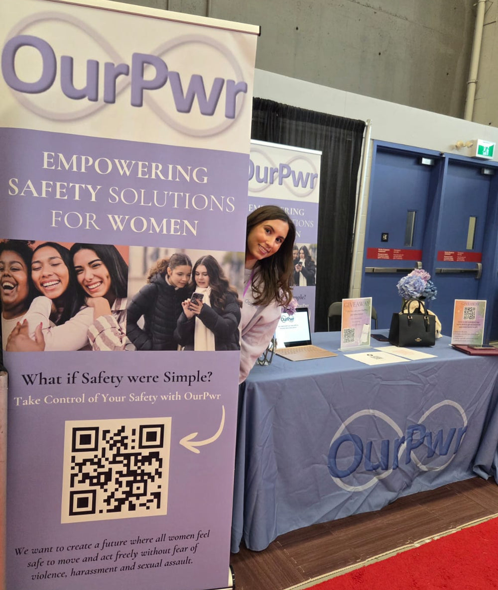 Robin Petrogiannis devant le stand d'OurPwr lors d'un événement, à côté d'une bannière déroulante où l'on peut lire "Des solutions de sécurité qui donnent du pouvoir aux femmes", avec des cartes promotionnelles et un ordinateur portable sur la table devant elle.